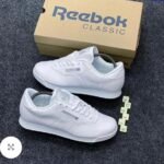 A pair of pristine white Reebok Classic sneakers with their matching box and Scrabble tiles spelling out 'REEBOK'.