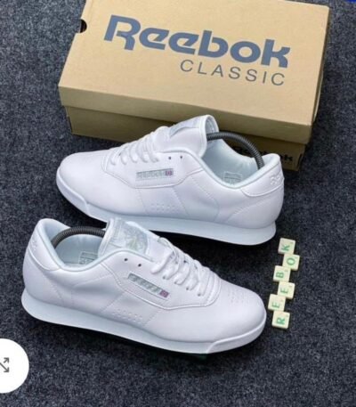 A pair of pristine white Reebok Classic sneakers with their matching box and Scrabble tiles spelling out 'REEBOK'.