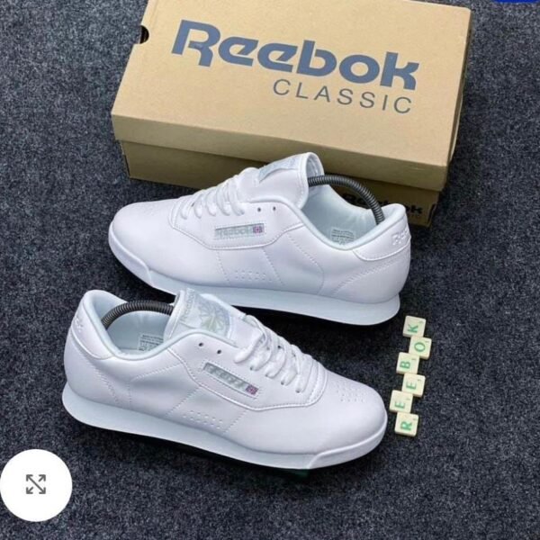 A pair of pristine white Reebok Classic sneakers with their matching box and Scrabble tiles spelling out 'REEBOK'.