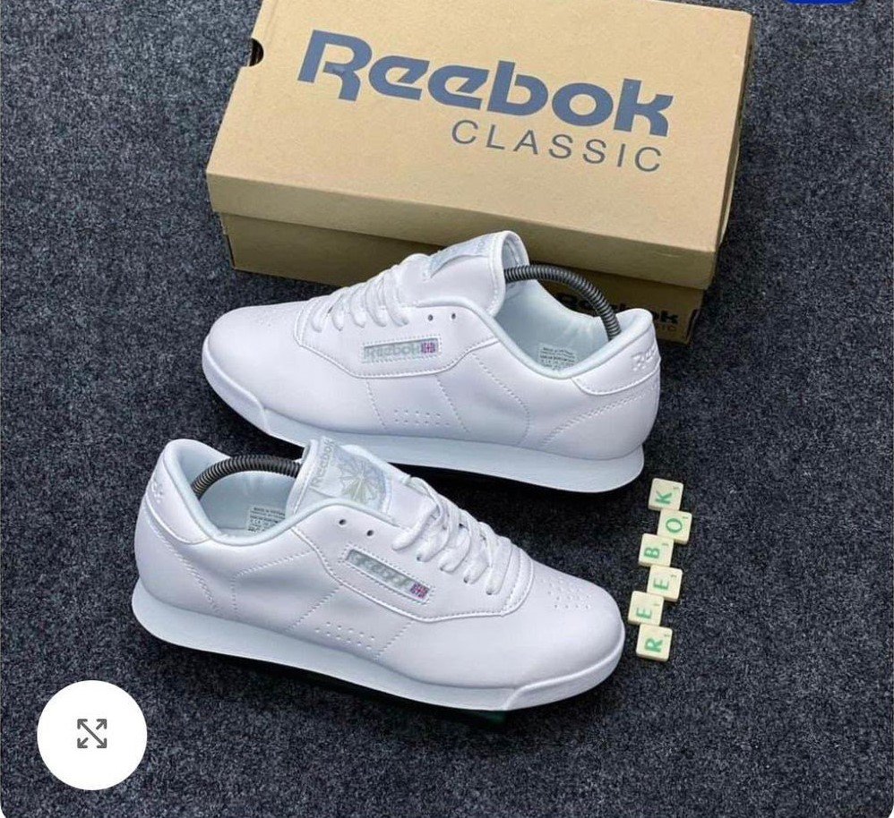 root A pair of pristine white Reebok Classic sneakers with their matching box and Scrabble tiles spelling out 'REEBOK'.