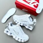 A pair of pristine white Nike sneakers with silver accents, accompanied by a red and white Nike shoe box and an extra pair of insoles, exuding luxury and style.