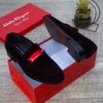 A pair of luxurious black Salvatore Ferragamo loafers with a distinctive red strap and gold buckle, presented in an open red shoe box.
