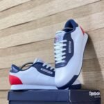 A pair of white, navy blue, and red Reebok Classic sneakers on a navy box against a wooden background.