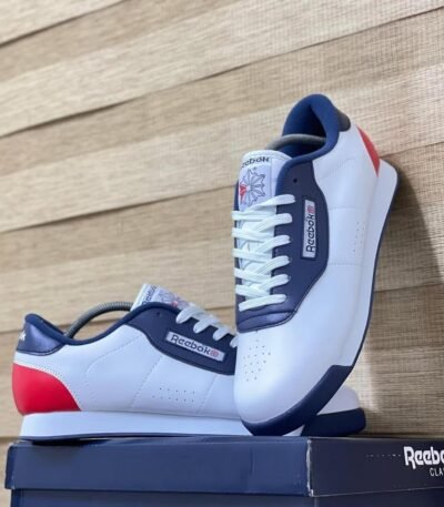 A pair of white, navy blue, and red Reebok Classic sneakers on a navy box against a wooden background.
