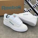 A pair of pristine white Reebok Classic sneakers with colorful accents, displayed with their box on a sleek grey surface.