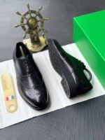 A pair of luxurious black leather oxford shoes with brogue detailing, presented on a crisp white background with a green box and golden accents, evoking a sense of high-end style and sophistication.