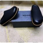 A pair of black leather mules with tan soles, positioned on either side of a Timberland shoe box, showcasing the shoes' sleek design and premium materials.