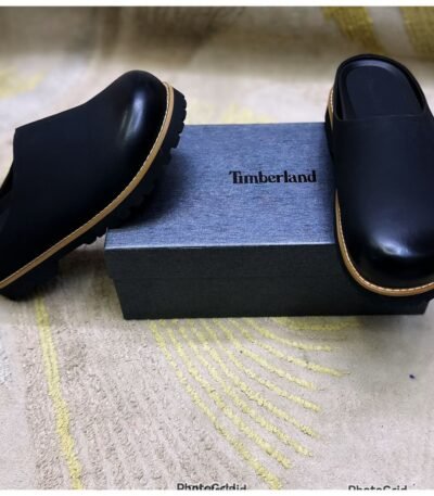 A pair of black leather mules with tan soles, positioned on either side of a Timberland shoe box, showcasing the shoes' sleek design and premium materials.
