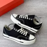 A pair of black sneakers with white laces and soles, placed on a grey wooden floor next to a red shoe box.