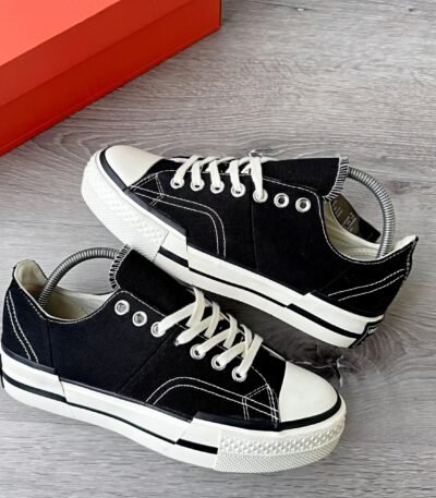 A pair of black sneakers with white laces and soles, placed on a grey wooden floor next to a red shoe box.