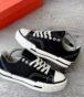 A pair of black sneakers with white laces and soles, placed on a grey wooden floor next to a red shoe box.
