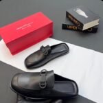 A pair of luxurious black leather half shoes with a sleek buckle, accompanied by a red shoe box and two fashion books, set against a sophisticated gray and white background.