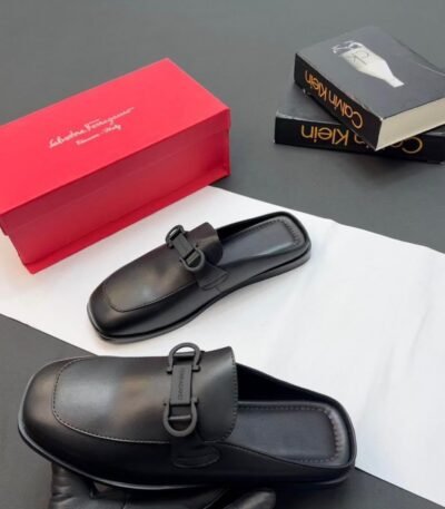 A pair of luxurious black leather half shoes with a sleek buckle, accompanied by a red shoe box and two fashion books, set against a sophisticated gray and white background.