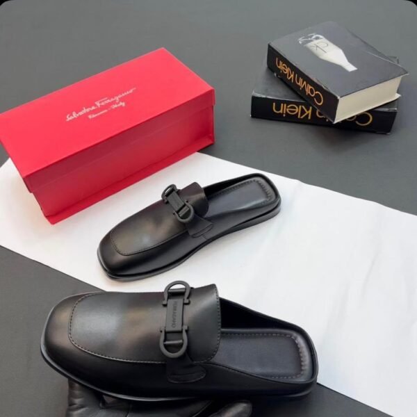 A pair of luxurious black leather half shoes with a sleek buckle, accompanied by a red shoe box and two fashion books, set against a sophisticated gray and white background.
