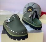 A pair of luxurious green half shoes with metallic studs and a silver buckle, showcased on a cream-colored shoe box.