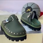 A pair of luxurious green half shoes with metallic studs and a silver buckle, showcased on a cream-colored shoe box.