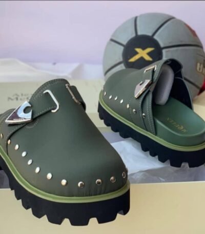 A pair of luxurious green half shoes with metallic studs and a silver buckle, showcased on a cream-colored shoe box.