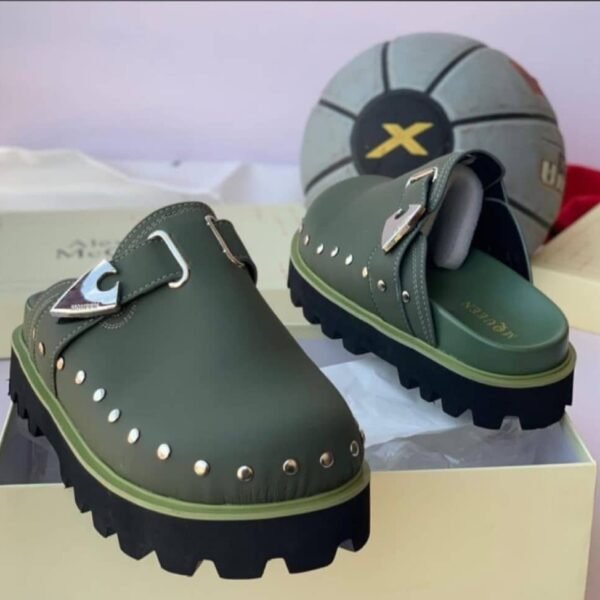 A pair of luxurious green half shoes with metallic studs and a silver buckle, showcased on a cream-colored shoe box.