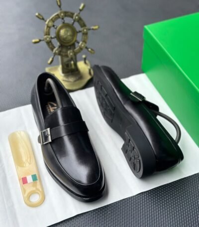 A pair of luxurious black leather loafers showcased on a pristine white surface, accompanied by a sleek shoe tree and a vibrant green box, evoking a sense of high-end sophistication.